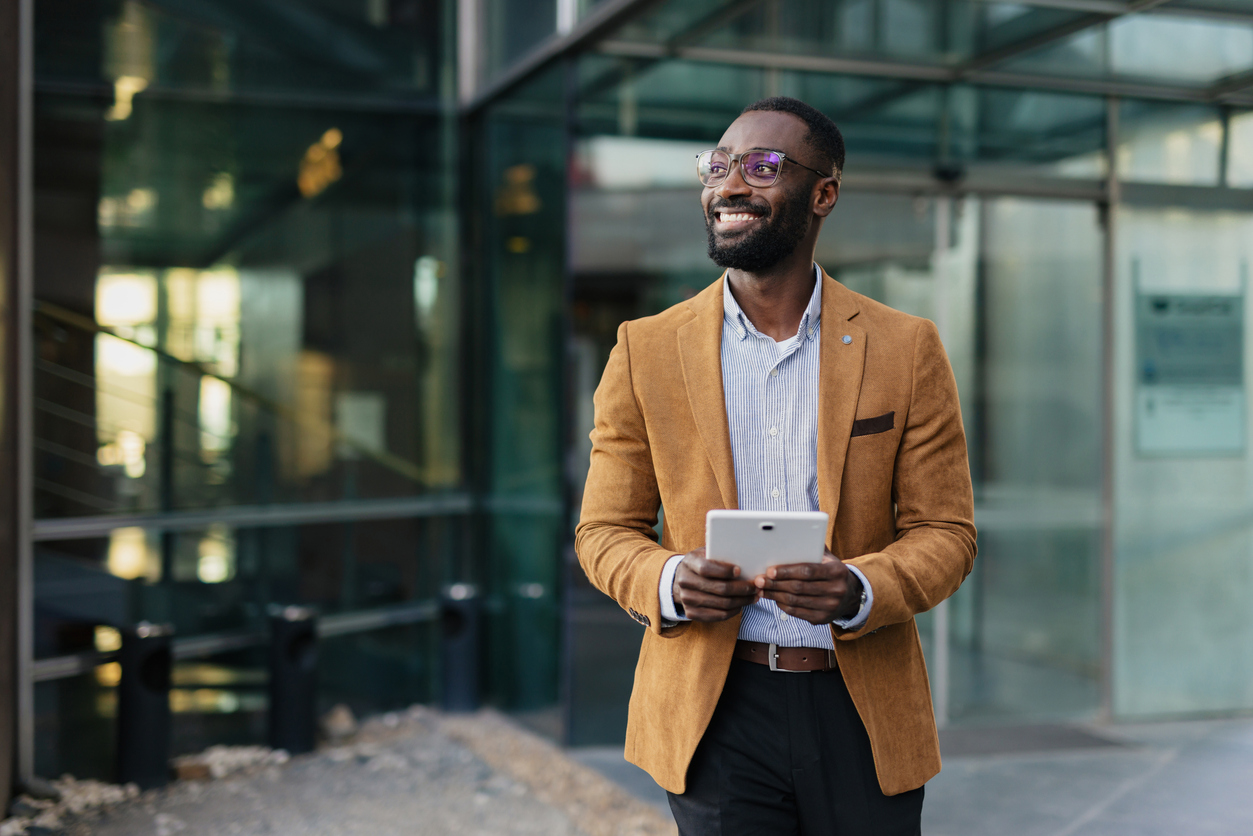 Mid adult businessman wearing glasses and a stylish jacket, confidently smiling while holding a tablet in an urban setting with modern architecture reflecting success